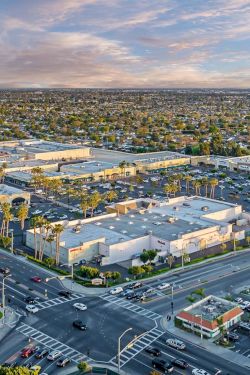 Aerial view of a suburban city at sunset with a large shopping center, busy roads, parking lots, and scattered palm trees.